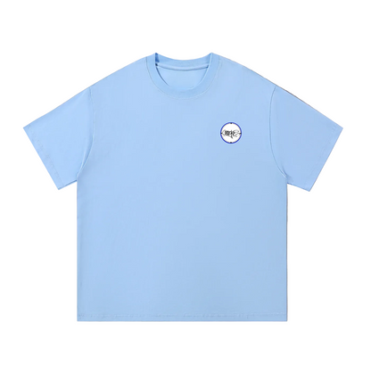 A light blue short-sleeve crewneck t-shirt with a small circular graphic on the left chest, laid flat on a textured gray background.